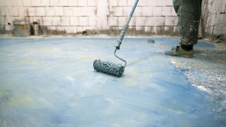 A person paints a concrete floor blue in a basement