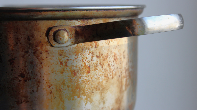 Closeup of a stained stainless steel pot with side handle; rust, heat marks, and heavy kitchen wear.