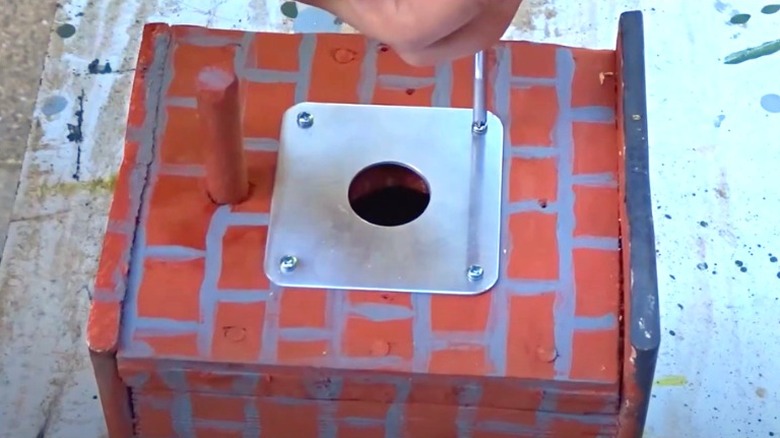 Person screwing metal cover plate onto brick-painted birdhouse