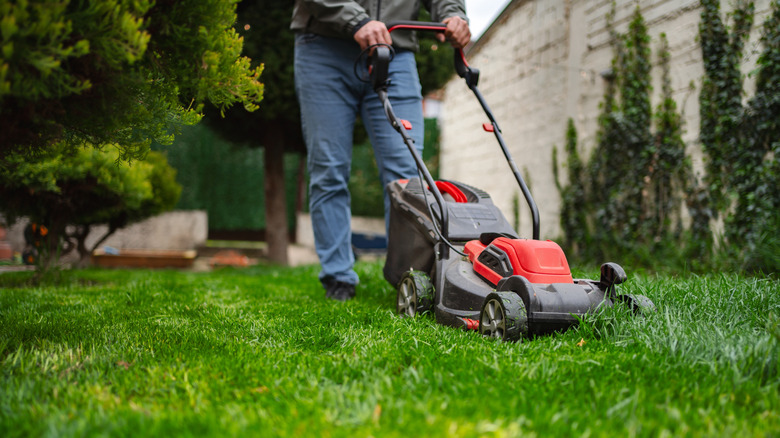 A person pictured from the waist down uses a lawn mower next to a large white building