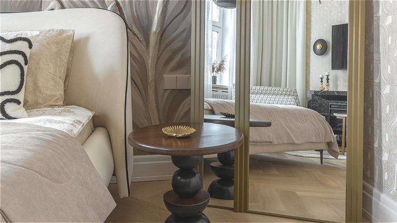 Bedroom corner with a mirror, wood table, gold dishes, and white bed.