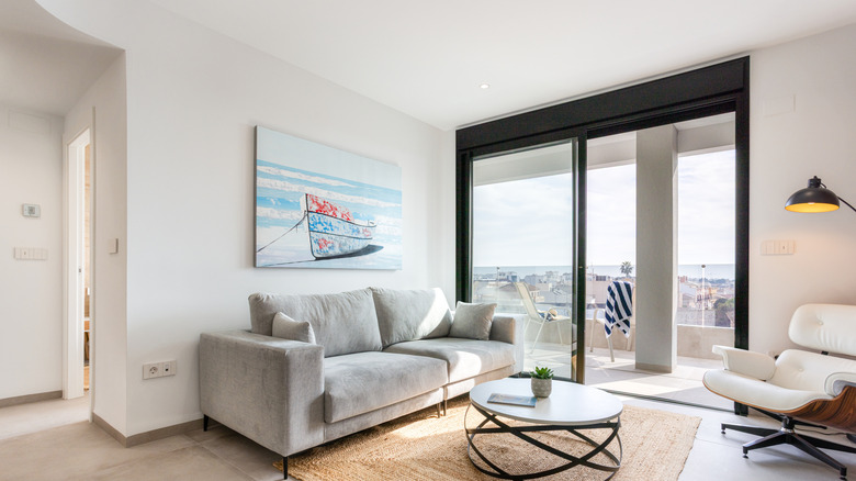 Bright minimalist living room featuring gray sofa, round coffee table, sliding glass doors to balcony, and soft coastal decor with sea view.