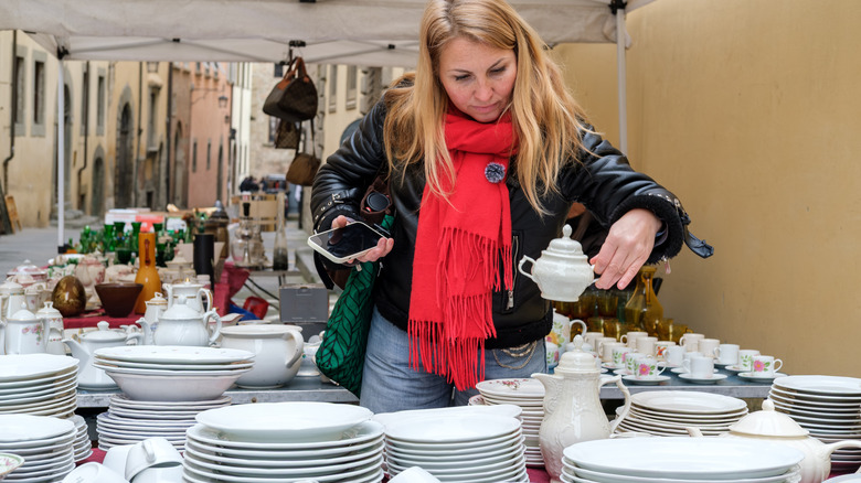 Blonde woman inspects a sugar bowl at a flea market at a table covered with various dishes.