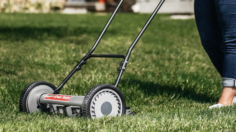A woman pushing a gray reel mower through green grass.
