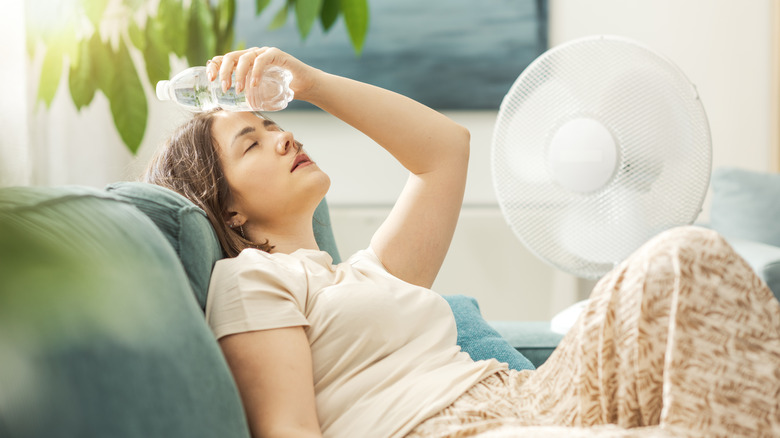 A young woman with a water bottle pressed to her forehead sitting in front of a fan.