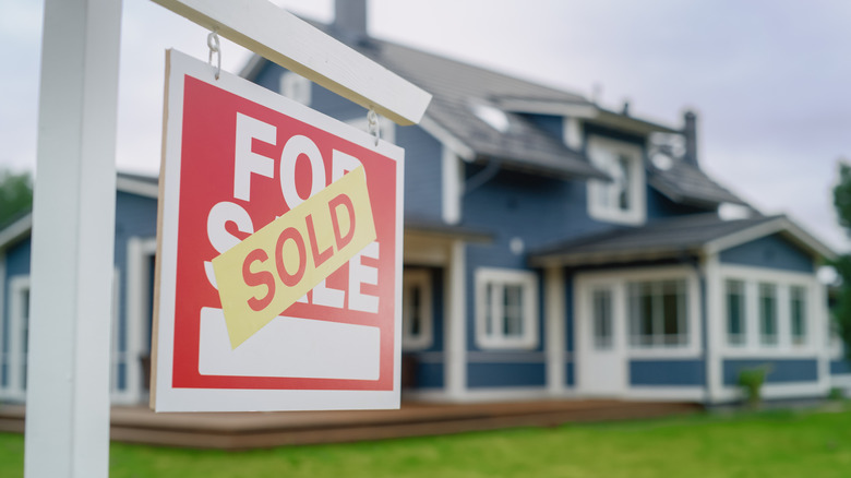 A red and white "for sale" sign updated with "sold" in front of a blurred blue house