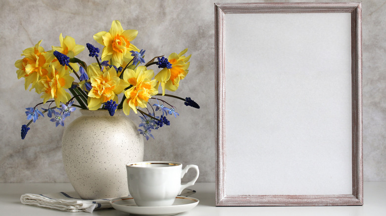 A simple spring floral arrangement with daffodils and hyacinth in a speckled white vase sit next to a cup and saucer and an empty photo frame.