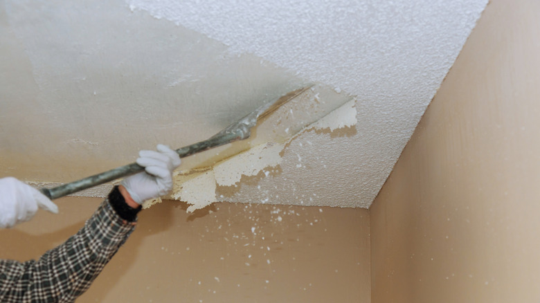 A person uses a scraper to remove a popcorn ceiling as debris falls