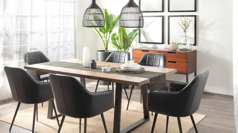 Black leather curved dining room chairs round a dining table with a stack of white plates, a white candle and table runner. There are two curved pendant lights hanging above the table.