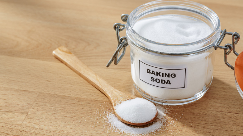 A open jar labeled "Baking Soda" on a wood table with a spoon overflowing with the powder.