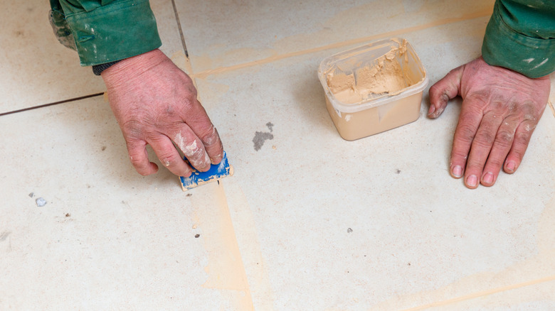 A man's hands are applying grout to tile with a small blue plastic tool.