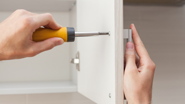 One hand uses a screwdriver to insert a screw into a white kitchen cabinet door while another holds a new handle on the other side of the cabinet door.