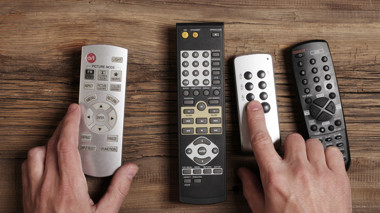 Person's hands holding two tv remotes, three more are on the table.