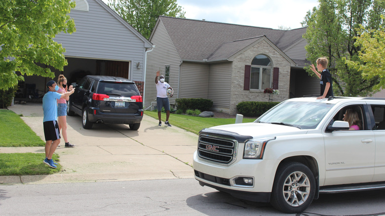 A group of people on the driveway of a home with a car in front of the garage and one driving past with people waving
