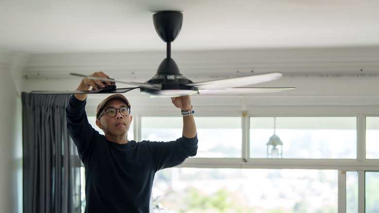 A man wearing a black shirt and glasses wipes the blades of a black ceiling fan.
