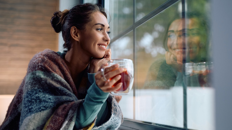 Woman wearing warm clothes, drinking a warm beverage, and looking out the window in the winter