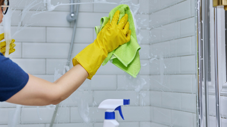 A person wearing yellow gloves and cleaning glass shower panels with a lime green cloth