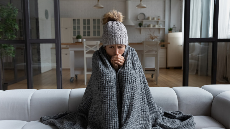 A woman sits on a couch bundled up with a hat and blanket in a cold house