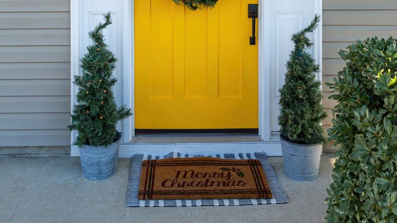 A "Merry Christmas" doormat sitting on a front porch in front of a yellow door