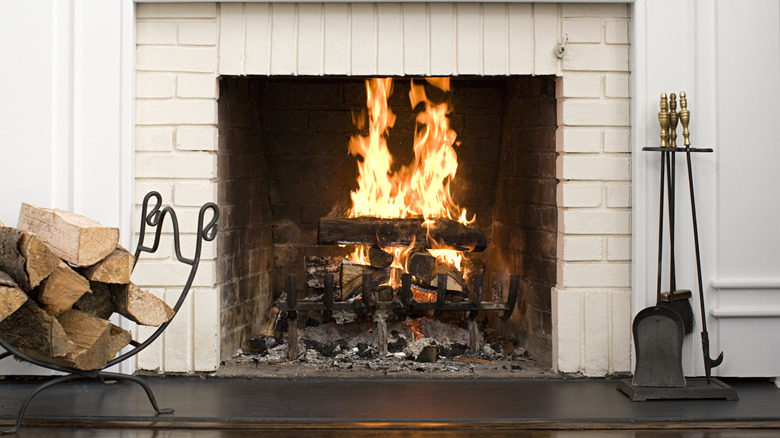 A fire burning in a fireplace in a house with some wood stacked on the left and fireplace tools on the right