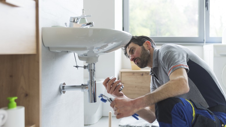 A professional plumber works underneath the white porcelain sink in a sleek, modern bathroom, replacing crucial components that require upkeep.