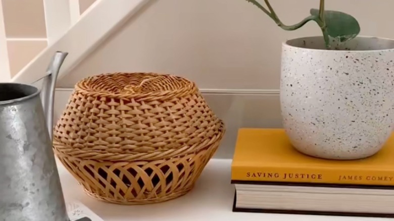 Two wicker baskets stacked by a staircase next to books, potted plant, and watering can