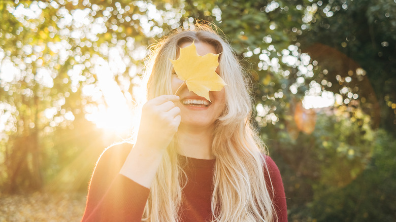 A smiling person holding a fall leaf over her face in the woods