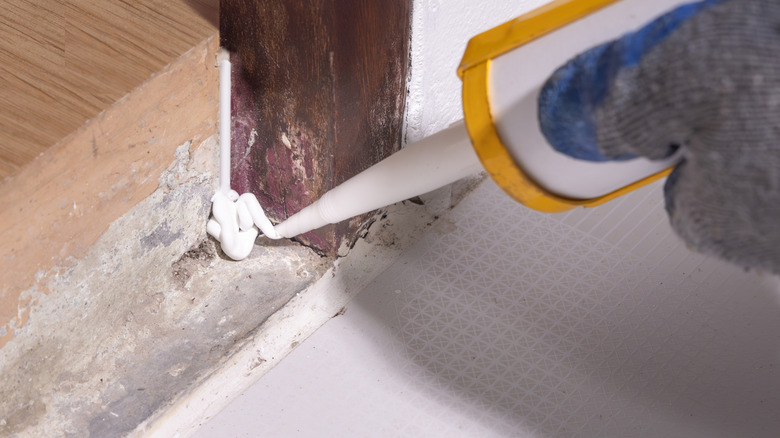A person caulks a gap in the wall of a home with a caulking gun and white caulk.