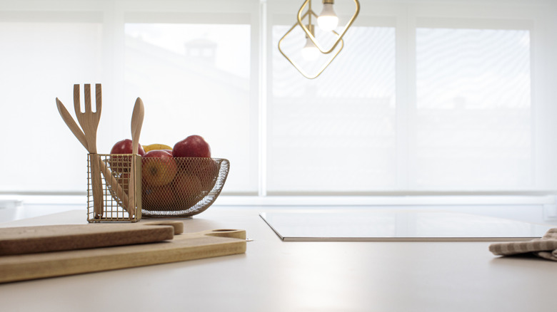 White kitchen counter with bowl of apples, wooden cutting boards, and wooden utensils.