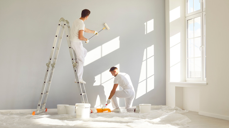 A pair of painters in all-white outfits paint a wall an off-white color in front of a window