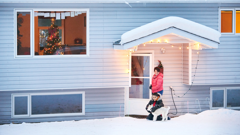 A woman, a child, and her dog walking into a house with basement windows in winter.