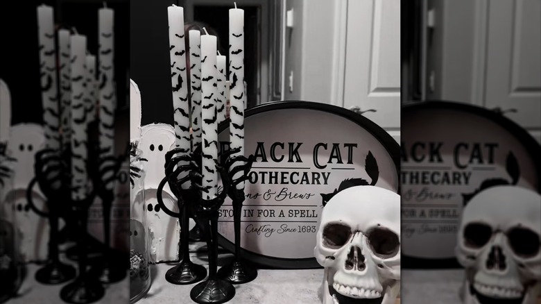 Set of black and white candles printed with bats inside of a skeleton candle holder next to a wooden black cat apothecary sign