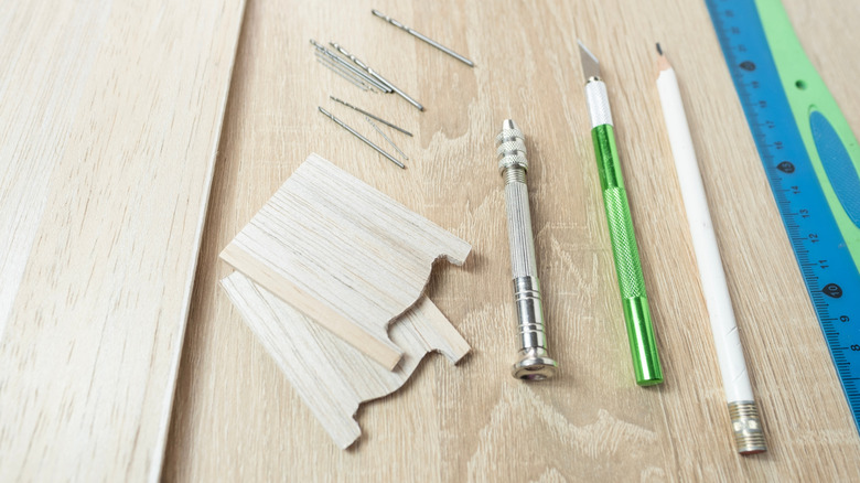 An exacto knife, a pencil, drill bits, and a ruler sitting on a sheet of balsa wood with two smaller pieces of balsa wood on top to do a DIY project
