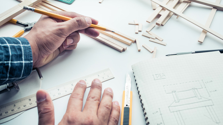 A man's hands holding a pencil drafting a plan for a chair with small pieces of balsa wood on the table around him