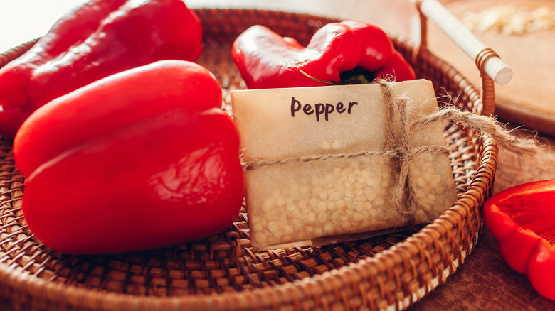 Pepper seeds in a paper envelope on a woven tray with fresh red peppers.