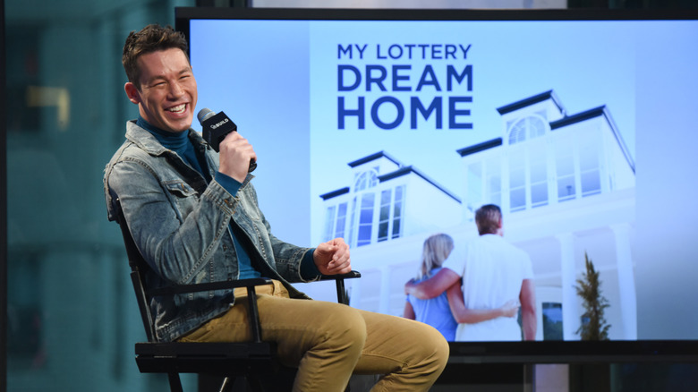David Bromstad smiles with a microphone in front of a My Lottery Dream Home display