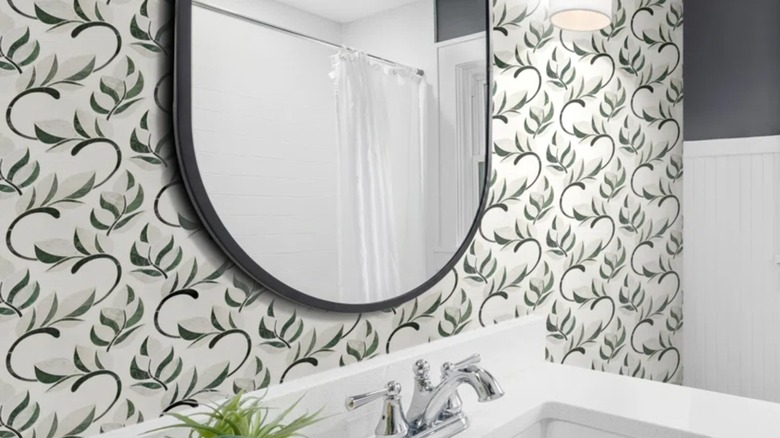 Partial view of a bathroom with waterjet mosaic green and black flower wall tiles, mirror with black frame, and white sink with chrome faucet.