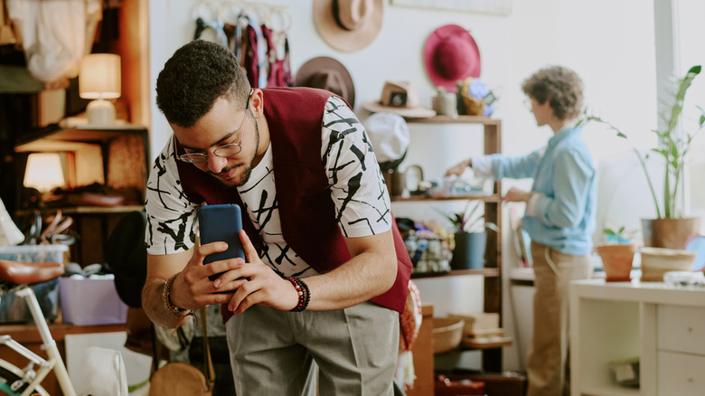 A shopper in a thrift or vintage store takes a photo of an object with their cellphone and another shopper looks at items on a shelf.