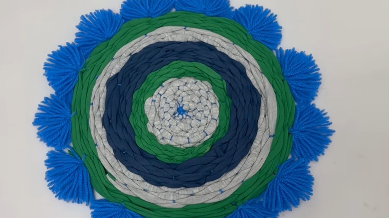 A woven blue, green, and gray mat made from t-shirts edged with blue yarn pom poms