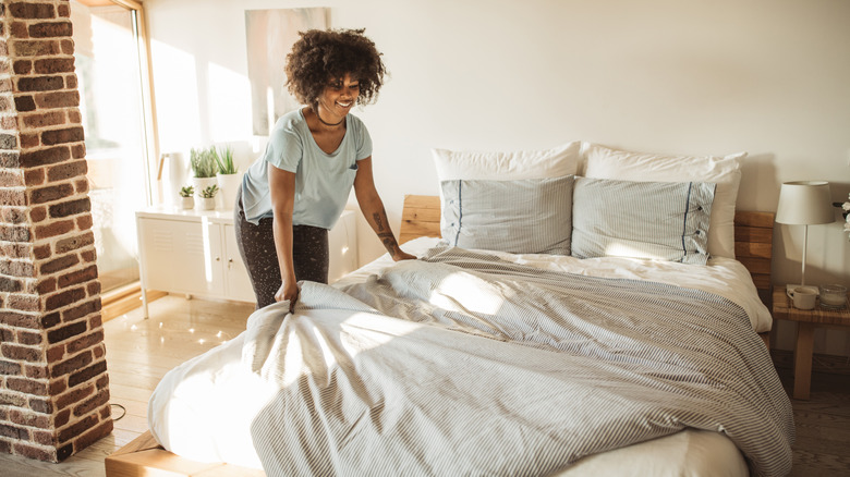 Woman making bed with soft decorative duvet and pillows in sunny bedroom