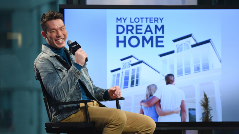 David Bromstad on stage smiling with a mic and a picture in the background that reads "My Lottery Dream Home"