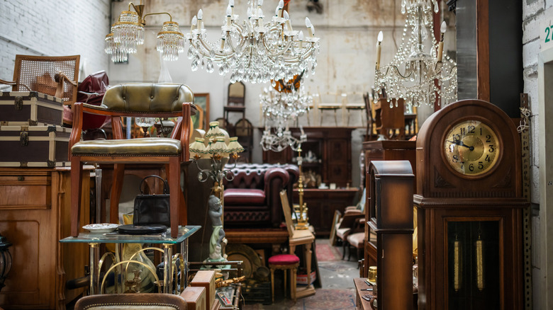 Store display with antique furniture stacked on top of each other, chandeliers, grandfather clock, tufted sofas, wooden chairs.