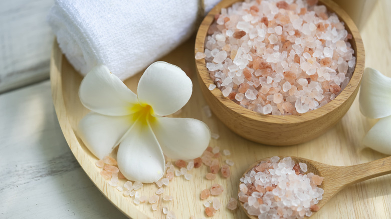Rock salt crystals in a wooden bowl with a spoon, flowers, and a rolled up towel