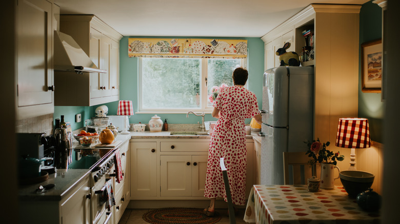 A woman in a red and white dress standing in front of a sink in a small, traditional style kitchen.