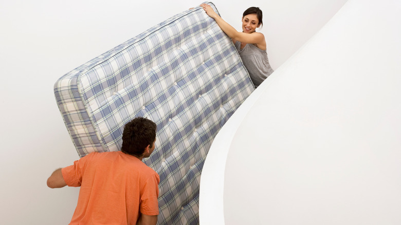 Man in orange shirt and woman in gray tank top carrying plaid mattress up white stairs with white wall