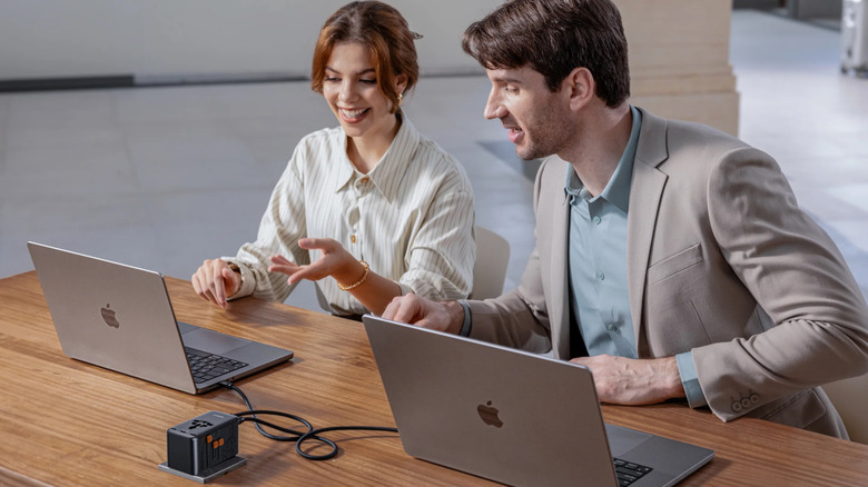 A man and woman sit next to each other at a desk while simultaneously charging their MacBooks with a Tessan Voyager 205 Universal Travel Charger