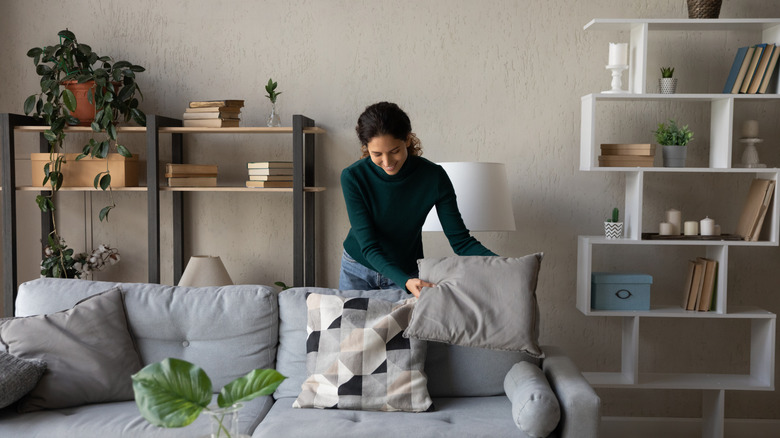 Woman fluffing pillows on living room couch with shelves in background
