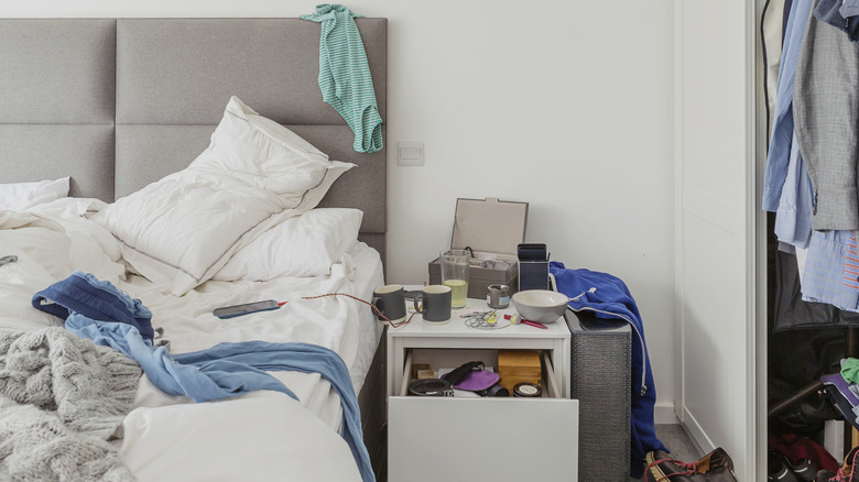 Messy bedroom with covered nightstand, plates, jewelry box, phone charger, etc.