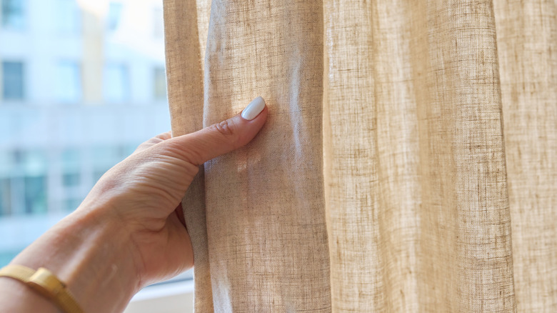 A woman's hand touching a beige linen curtain in front of a sunny window