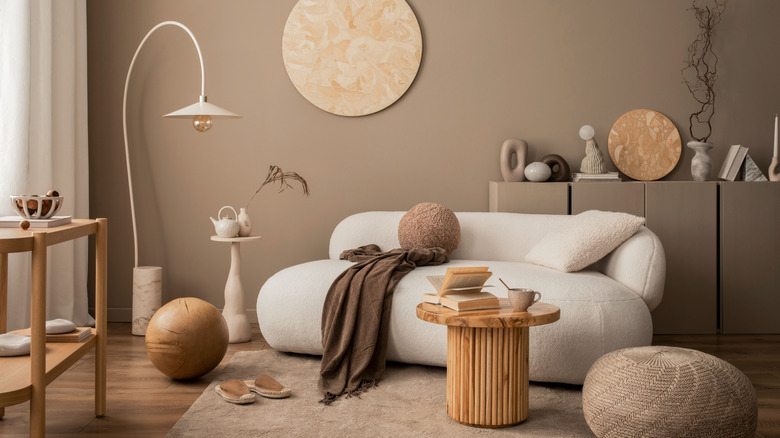Interior design of a cozy living room with wooden wall panel, stylish sofa, round coffee table, braided pouf, brown sideboard, vase with branch, beige lamp and personal accessories.